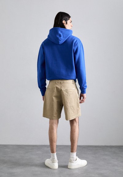 Felpa blu accorciata, pantaloni beige, sneakers bianche; mostra un taglio rilassato con tasca a marsupio e cappuccio, tessuto texturizzato e design informale.