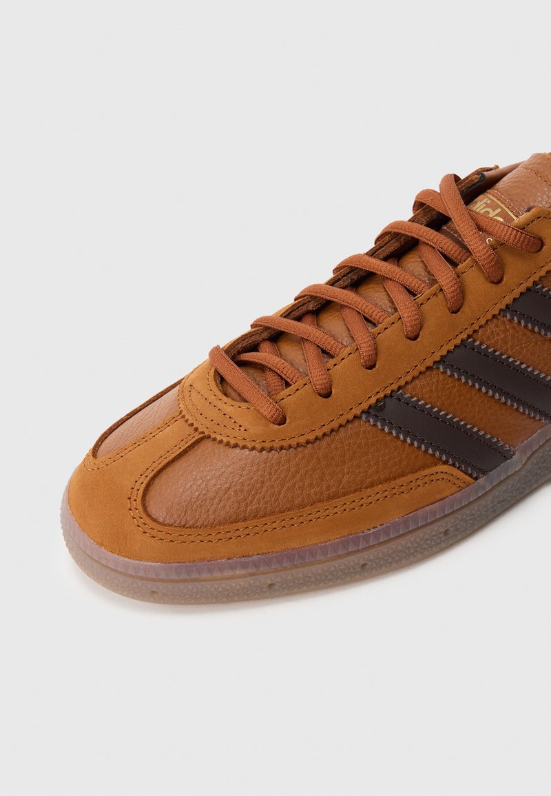 Brown leather sneakers featuring three dark brown stripes, textured finish, round toe, and brown laces. Clear rubber sole with traction pattern.
