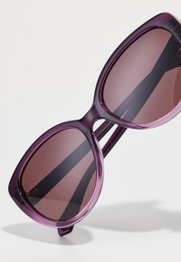 Lunettes de soleil en forme d'œil de chat violettes avec des verres dégradés bruns. Fabriquées en plastique lisse, avec une fine monture et des branches larges.