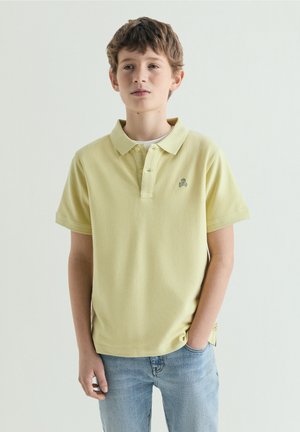Dreng med kort brunt hår iført en lysegul polo-shirt og lyseblå jeans, står mod en ensfarvet baggrund.