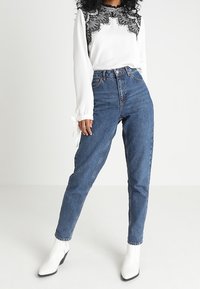 Blouse blanche avec détails en dentelle noire aux épaules, associée à un jean en denim bleu taille haute et des bottines blanches.