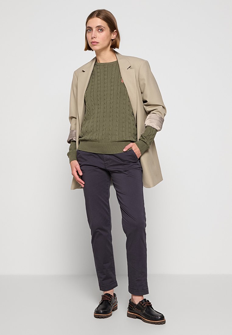 Beige Blazer über olivgrünem Kabelstrickpullover, kombiniert mit dunklen Hosen und schwarzen Schuhen. Einfaches Design mit strukturierten Materialien und gedämpften Farben.