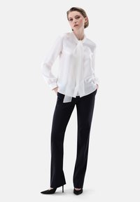 Blusa bianca trasparente con dettaglio del collo annodato, maniche lunghe con polsini arricciati, abbinata a pantaloni neri aderenti e décolleté neri a punta.