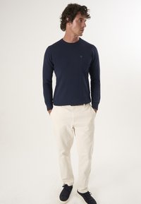 Marineblauw longsleeve shirt met geribbelde manchetten, gedragen met crèmekleurige broek en zwarte sneakers. Bevat een klein groen logo op het shirt.