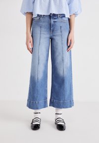 Jeans a gamba larga blu chiaro con vita alta, caratterizzati da dettagli di cucitura frontale e orlo piegato. Abbinati a scarpe nere e calzini bianchi.