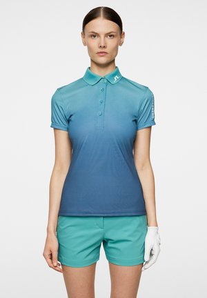 Una polo con gradiente in tonalità teal e blu, a maniche corte con colletto, dotata di bottoni e logo sulla spalla, abbinata a pantaloni corti teal.