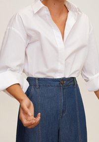 Chemise en coton blanc à boutons avec des poignets retroussés, associée à un pantalon en denim bleu taille haute avec un bouton à l'avant et des détails cousus.