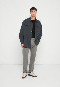 Giacca camicia grigio scuro con due tasche sul petto, indossata sopra un top nero, abbinata a pantaloni grigio chiaro e sneakers color crema. Texture liscia.