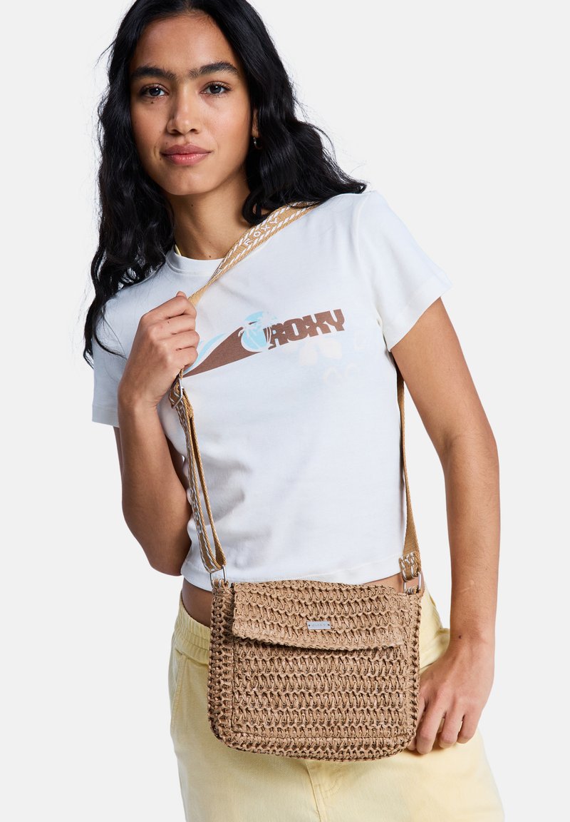 Młoda kobieta o ciemnych włosach nosi biały t-shirt z nadrukiem i jasnżółte spodnie, trzyma beżową plecioną torbę na ramię.