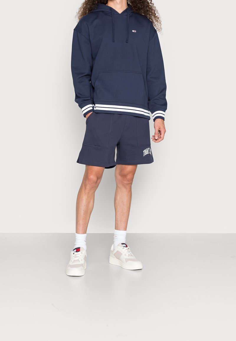 Marinblå hoodie med vita randiga ärmslut och midja, kombinerad med matchande shorts med en logotyp, bärs med ljusa sneakers och vita strumpor.