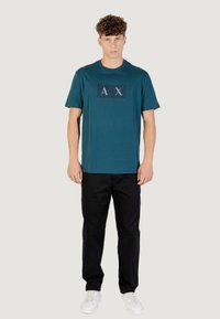 T-shirt en coton teal avec un col rond et des manches courtes, arborant un graphique de logo en bleu foncé et blanc. Associé à un pantalon chino noir et des baskets blanches.