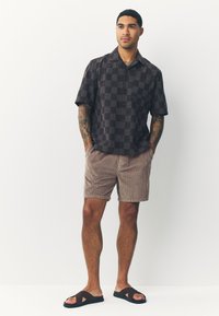 Kurzärmeliges kariertes Hemd in dunklen Tönen, kombiniert mit hellbraunen Cord-Shorts und braunen Pantoletten. Durchweg glatte Textur.