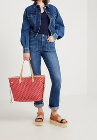 Chaqueta de denim y jeans combinados con un bolso tote rojo con estampado y sandalias de plataforma marrones. El conjunto enfatiza un estilo casual y funcional.