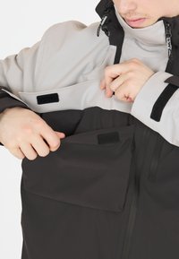 Veste imperméable avec des panneaux gris et noirs, dotée de poches avec Velcro, de poignets ajustables et d'un col haut pour une protection optimale.