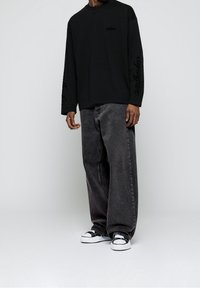 Musta pitkähihaisen paidan rintaan ja hihaan painettu logo; yhdistetty löysälahkeisiin tummanharmaisiin farkkuihin ja mustavalkoisiin tennareihin.