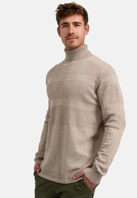 Beige, magas nyakú pulóver texturált csíkokkal, hosszú ujjakkal és bordázott mandzsettával. Simább anyag, laza kialakítással, zöld nadrággal párosítva.