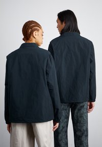 Deux personnes font dos, vêtues de vestes légères noires à la conception simple. La veste présente des poignets élastiques et une coupe décontractée.