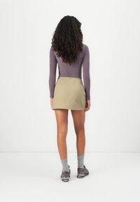 Person with long curly hair stands facing a white wall wearing a purple long-sleeve top, beige skirt, grey socks, and dark sneakers.