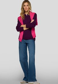 Lila langärmeliges Oberteil mit einem pinkfarbenen Pullover über den Schultern, kombiniert mit ausgestellten blauen Jeans und metallisch glänzenden Schuhen.