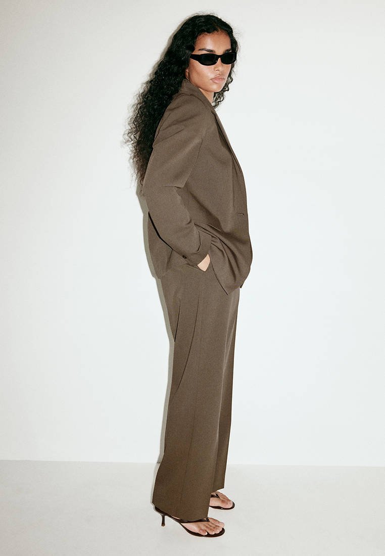 Femme portant un costume marron oversize et des lunettes de soleil noires, debout de côté avec les mains dans les poches et portant des sandales noires à talons.