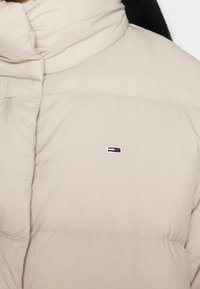 Beige puffjacka med hög krage, kviltad textur och distinkt logotyp med horisontella ränder i rött, vitt och blått.