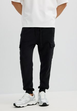 Persona con pantalones jogger cargo negros y zapatillas deportivas blancas, de pie contra un fondo claro y liso.