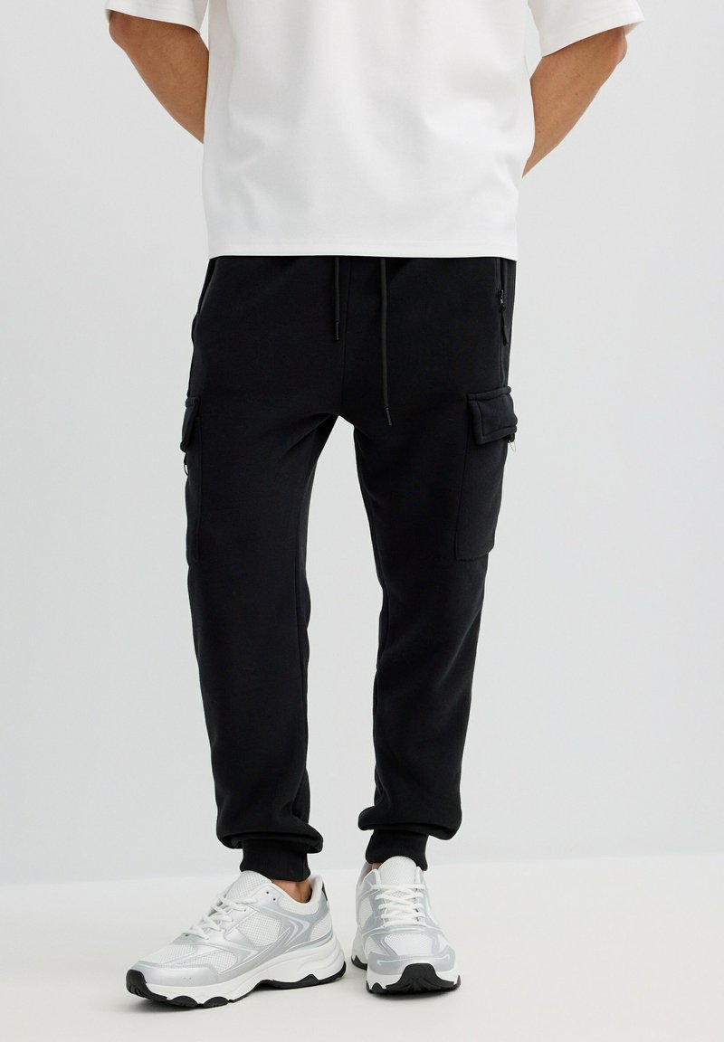 Persona con pantalones jogger cargo negros y zapatillas deportivas blancas, de pie contra un fondo claro y liso.