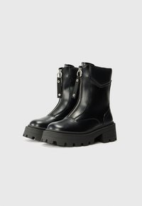 Botas de cuero negras hasta el tobillo con suelas gruesas y de gran tracción, y cremalleras frontales expuestas con lengüetas de tela.