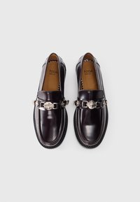 Loafers din piele maro închis cu un accent decorativ dintr-un lanț argintiu și detalii de cusătură, având un vârf rotunjit și o talpă robustă din cauciuc.