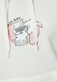Sudadera blanca con un gráfico de un gato tumbado sobre su espalda con un portátil. El gato tiene pelaje gris y blanco, y el diseño incluye colores pastel.