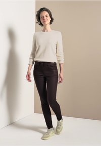 Donkerbruine skinny jeans, lichtbeige lange mouw top. Zachte textuur, eenvoudig ontwerp. Lichte sneakers met gele accenten. Model staat tegen een neutrale achtergrond.