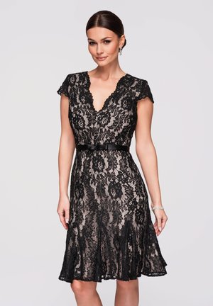 Frau in einem knielangen schwarzen Spitzenkleid mit kurzen Ärmeln, V-Ausschnitt, tailliert mit Gürtel und dezentem Blumenmuster, stehend vor schlichtem Hintergrund.