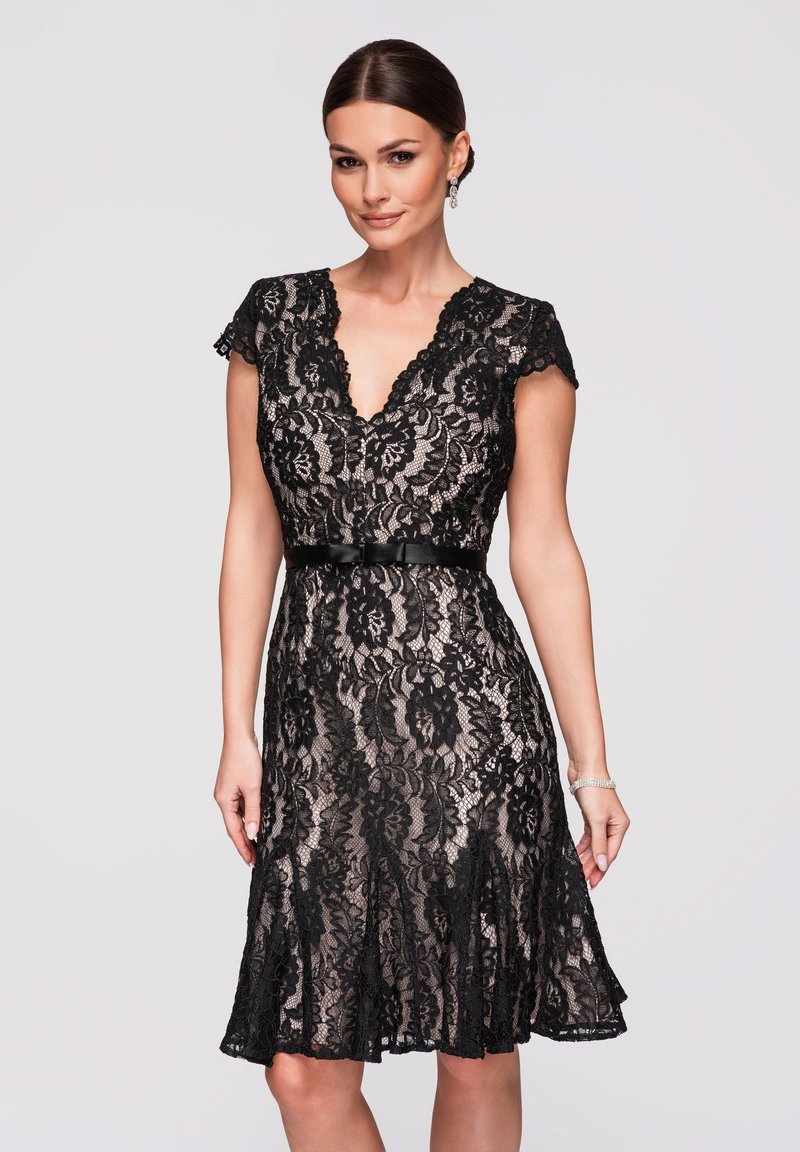 Femme portant une robe noire en dentelle mi-longue avec des manches courtes, un col en V, une taille ajustée avec une ceinture et un motif floral subtil, se tenant devant un fond uni.