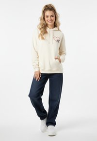 Felpa color crema con grafica a cuore rosso, tasca frontale, abbinata a jeans di denim blu scuro e sneakers bianche. Design semplice e vestibilità rilassata.