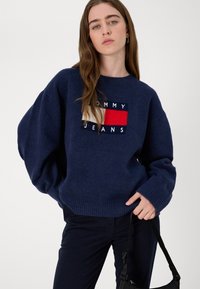 Maglione lavorato a maglia blu navy con vestibilità ampia, polsini a coste e una patch con logo nei colori rosso e bianco. Abbinato a pantaloni scuri e una borsa.