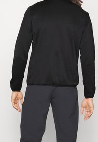 Mężczyzna w czarnej koszuli z długimi rękawami i ciemnoszarych spodniach, stojący z odprężonymi ramionami, pokazany od tyłu na jednolitym tle.