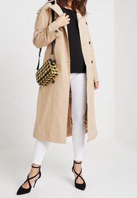 Cappotto trench beige con una silhouette lunga, abbinato a jeans skinny bianchi e tacchi neri con cinturini. L'outfit include una borsa a tracolla gialla a scacchi.