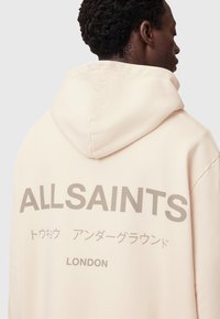 Felpa rosa chiaro con cappuccio regolabile, con grande scritta grigia "ALLSAINTS" e caratteri giapponesi sul retro. Texture morbida.