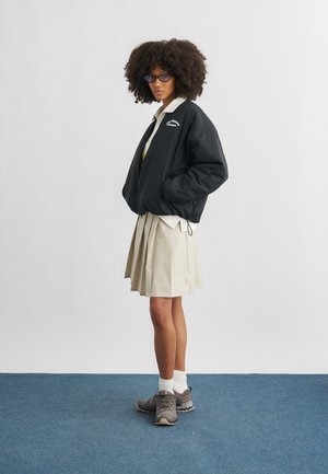 Femeie cu păr creț și ochelari, purtând o geacă neagră, fustă plisată bej, șosete albe și adidași gri, stând pe o mochetă albastră.