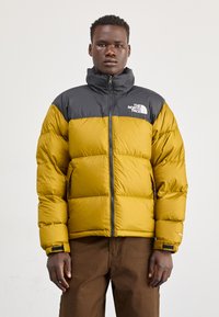 Heren pufferjack met een gele romp en een zwarte kraag. Heeft een rits aan de voorkant, een logo op de borst en een gewatteerde textuur.