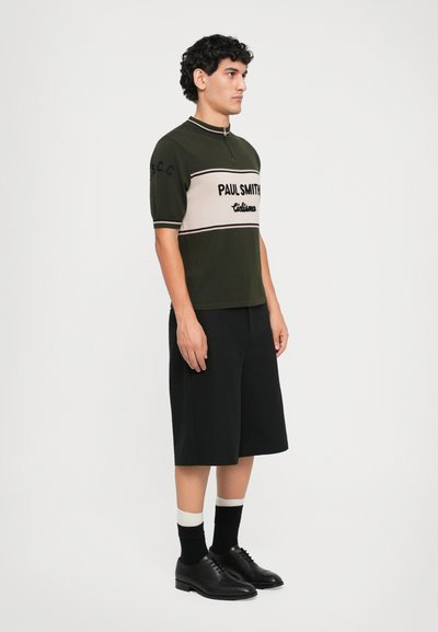 Olivengrünes Kurzarm-Strickoberteil mit beiger horizontaler Streifen und schwarzer Schrift; kombiniert mit schwarzen Shorts und schwarzen Schnürschuhen.