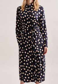 Vestido azul marino con mangas largas, cierre delantero de botones y cintura con lazo. Presenta grandes lunares blancos y naranjas y un bolsillo en el pecho.