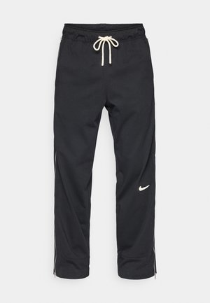Pantalons de jogging noirs avec une taille élastique et un cordon de serrage. Présente des zips latéraux et un logo Nike blanc sur la jambe gauche. Texture lisse.