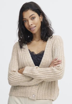 Hellbeige, gerippter Cardigan, strukturierter Strick, tiefer V-Ausschnitt, mit zwei großen, dekorativen Knöpfen. Trägt man über einem schwarzen Spitzenoberteil.