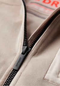 Beige suede jacket featuring a black zipper with "SUPERDRY" branding. Interior tag visible with red and gray text. Smooth texture, tailored design.