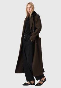 Abrigo largo de lana marrón con acentos de cuero negro en los hombros, combinado con un suéter negro texturizado y pantalones negros de pierna ancha, completado con zapatos negros planos.