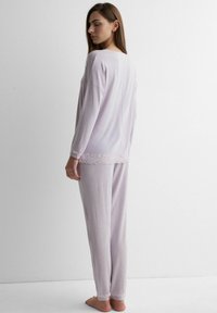 Frau steht barfuß seitlich, trägt ein helllila Langarmoberteil und eine Hose mit Spitze an den Bündchen und am Saum.