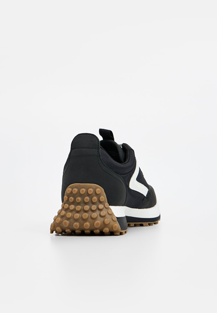 Chaussure de sport noire dotée d'une semelle en caoutchouc texturée, d'un dessus en tissu doux et d'un accent blanc sur le côté. Comprend une languette au talon.