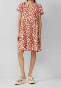 Donna che indossa un abito corallo a maniche corte e lunghezza ginocchio con macchie leopardate rosa chiaro e sandali con piattaforma beige.