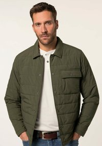 Olivgrüne Steppjacke mit Druckknöpfen, einer Brusttasche und Kragen, getragen über einem weißen Hemd und blauen Jeans.